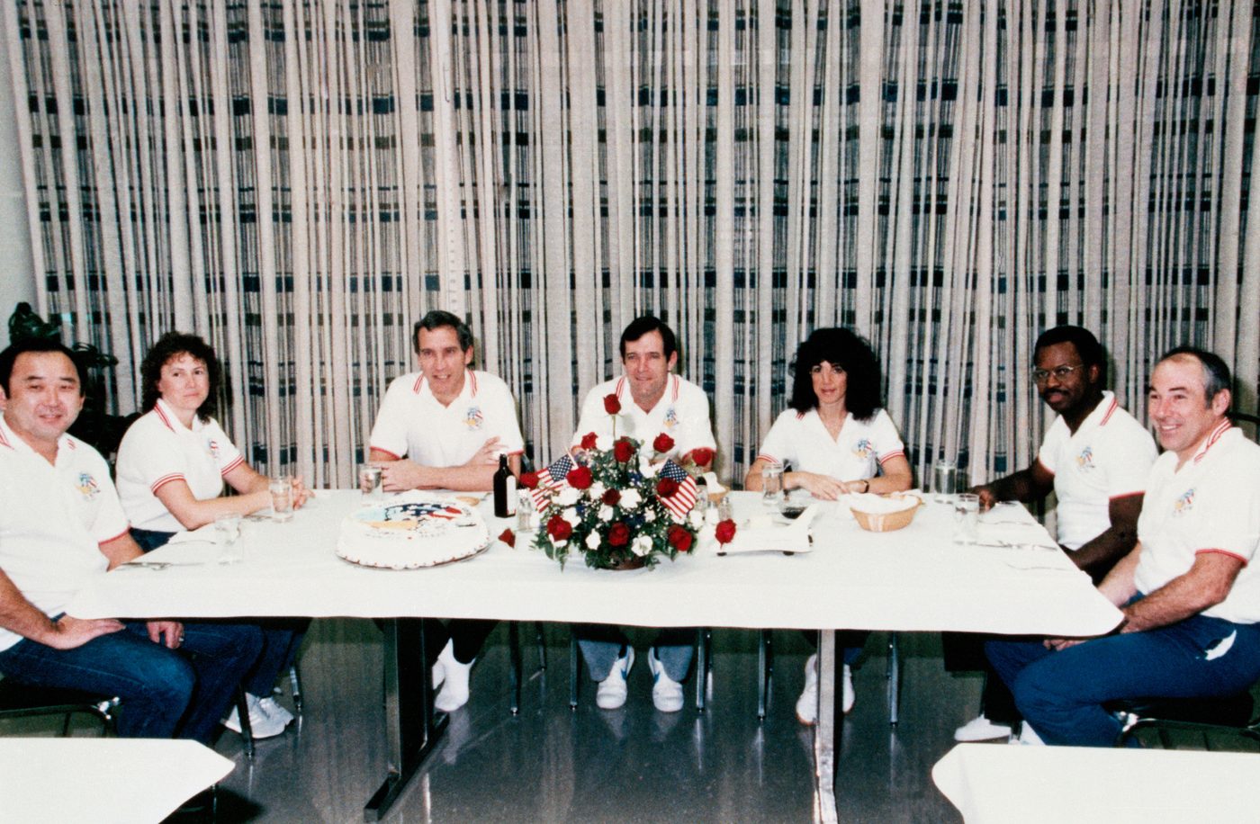 Os sete tripulantes da missão STS-51L sentados à mesa durante o café da manhã do dia do lançamento, no prédio de operações e verificação