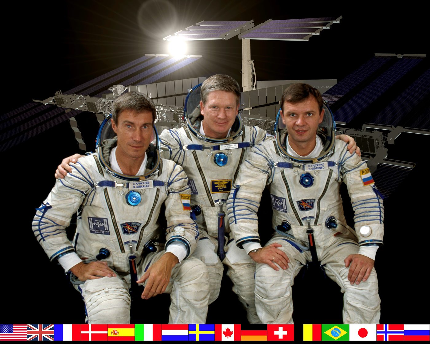 Retrato oficial da tripulação da Expedição 1, com Shepherd ao centro, Gidzenko à direita e Krikalev à esquerda, em trajes Sokol, diante de uma representação da ISS