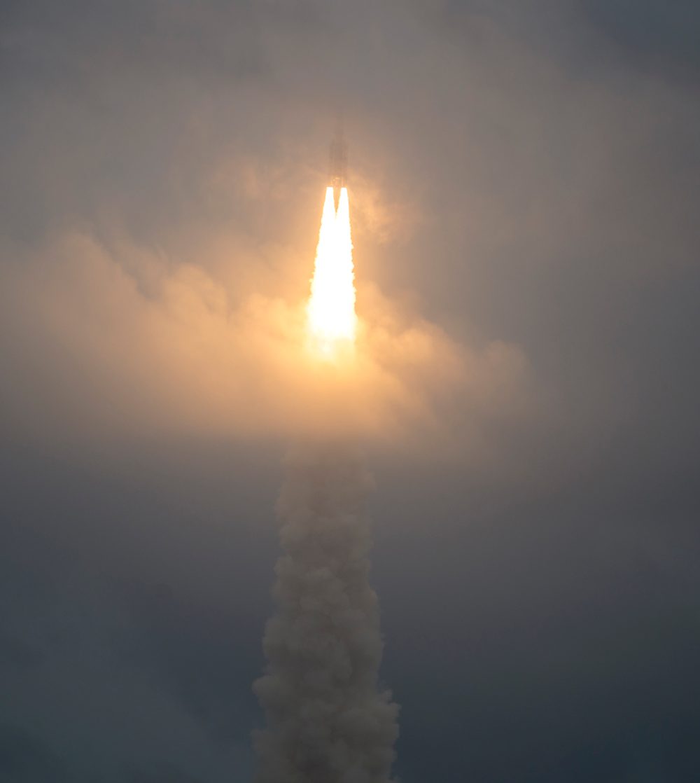 Foguete Ariane 5 em ascensão, com chama intensa e coluna de fumaça, atravessando nuvens no lançamento do telescópio Webb