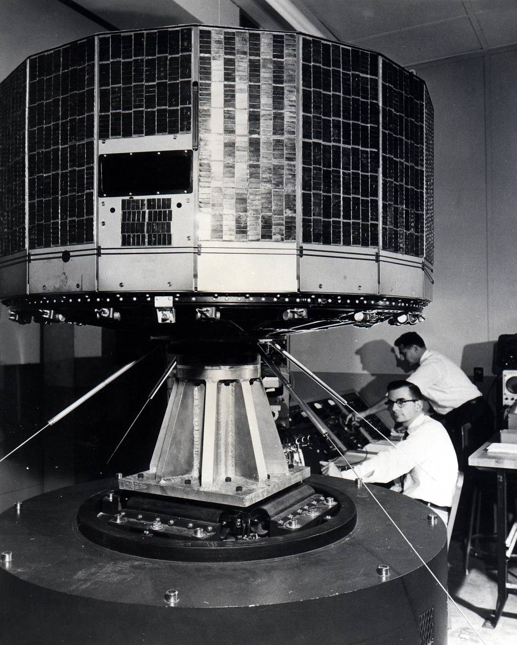 Fotografia em preto e branco do satélite TIROS 1, em forma de tambor, coberto por células solares, em teste de vibração, com dois técnicos ao fundo