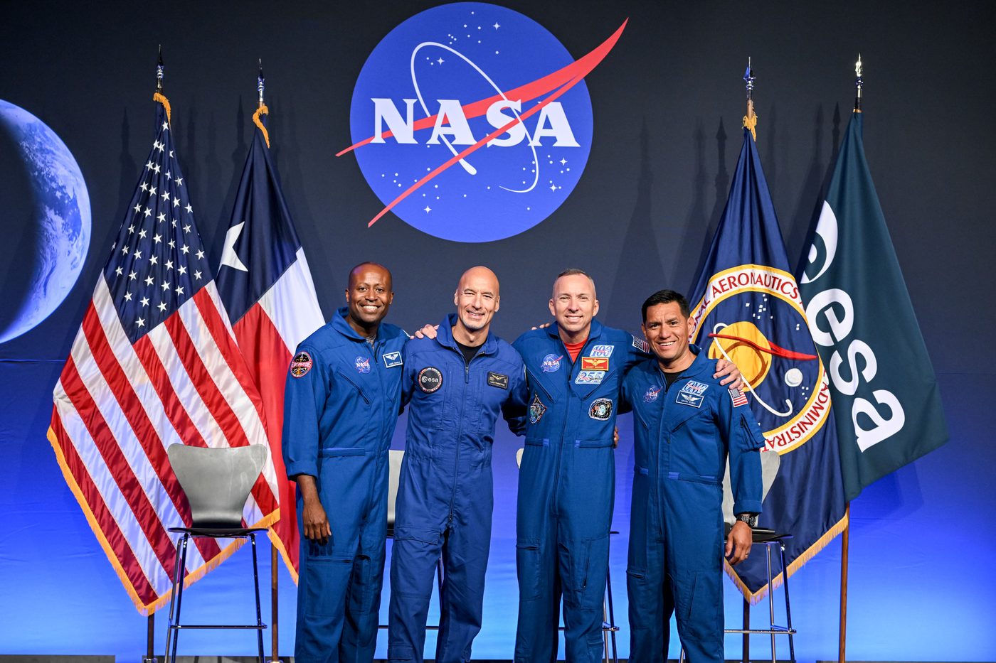 Os quatro astronautas da Artemis III posam lado a lado, de braços dados, diante de bandeiras dos Estados Unidos, do Texas, da NASA e da ESA.